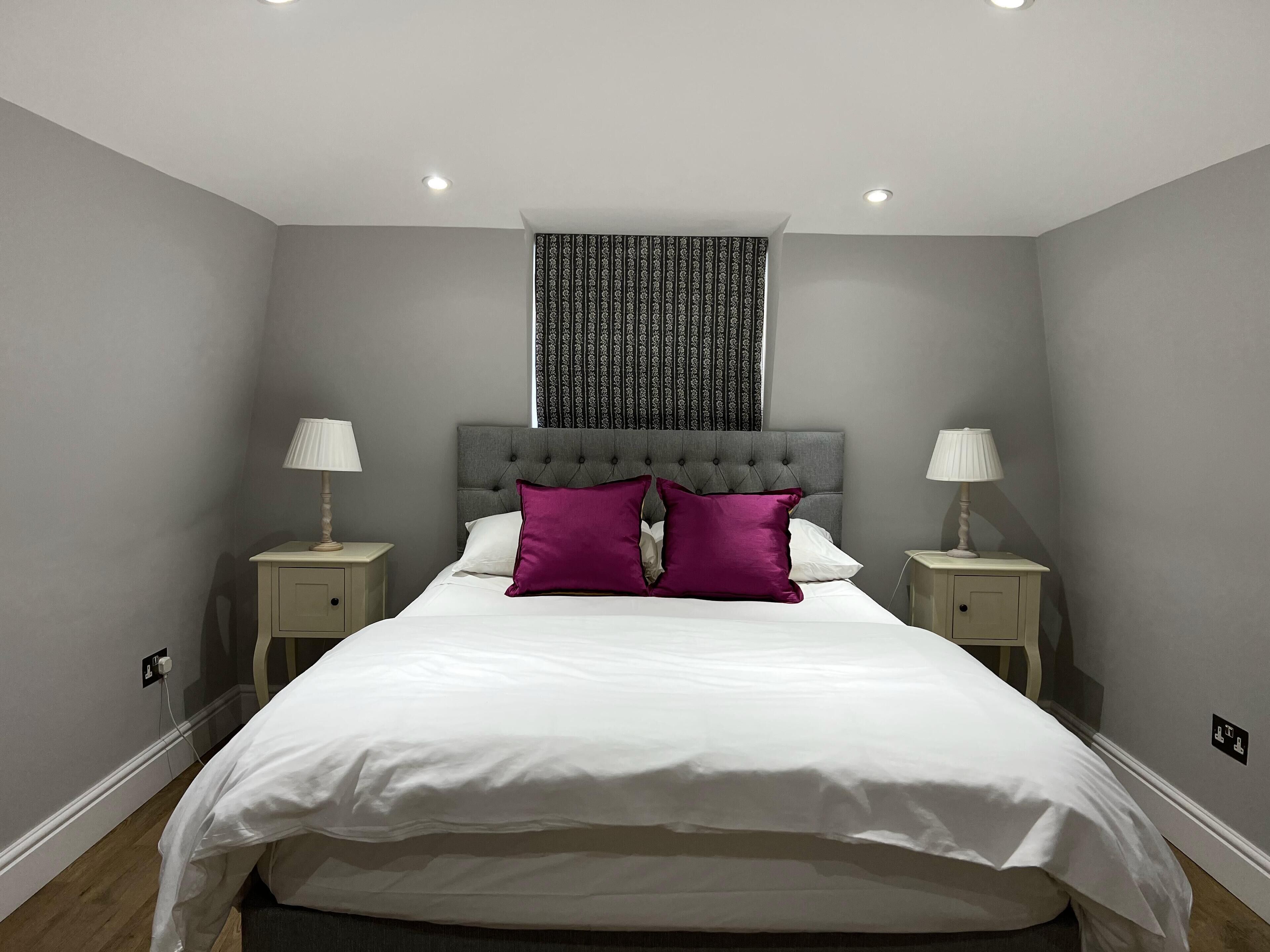 A neatly made bed with white linens and two purple pillows, flanked by two bedside tables with lamps, in a room with grey walls and a patterned curtain.