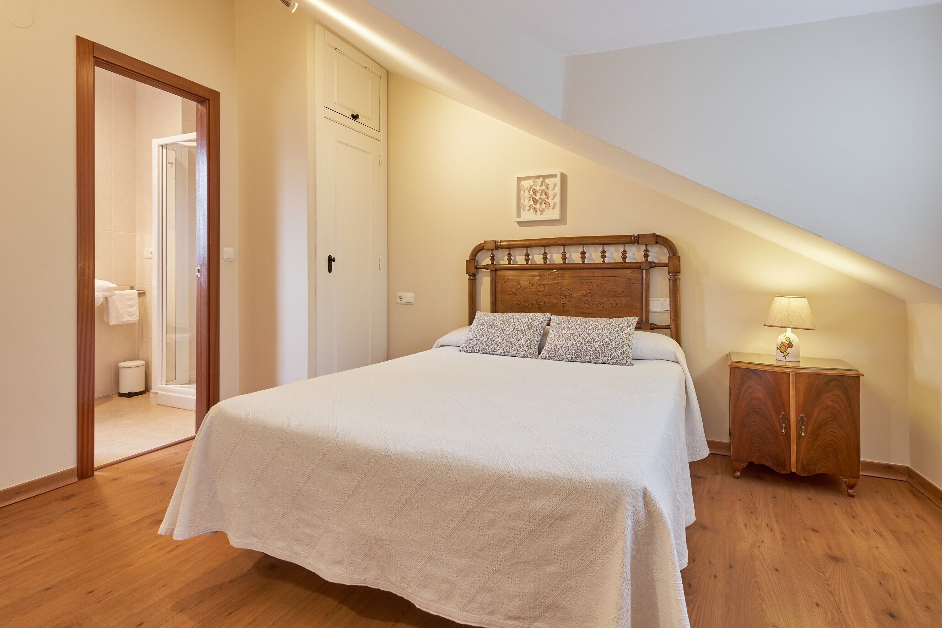 Une chambre à coucher comprenant un lit, une tête de lit en bois, une table de chevet, une lampe et une porte donnant sur une salle de bain.