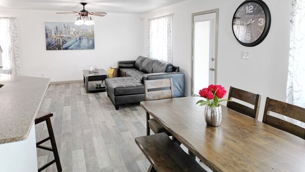 Ein modernes Wohnzimmer mit grauem Sofa, einem Esstisch mit Stühlen, einer Küchenzeile und einem Deckenventilator.