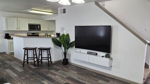 Eine moderne Küche mit weißen Schränken, einer Mikrowelle und einem großen Flachbildfernseher an der Wand. Zwei Holzschelle und ein Blumentopf sind ebenfalls vorhanden.