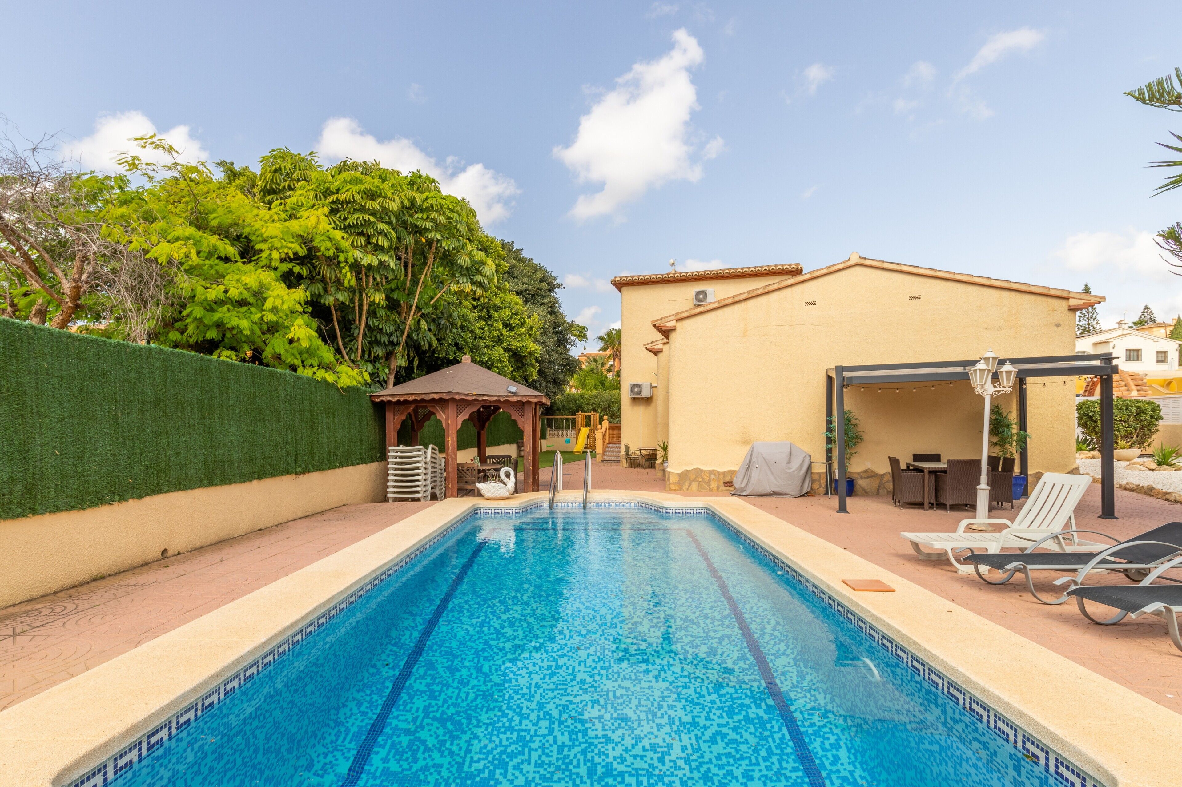 A swimming pool with lounge chairs, a gazebo, and a building with a tiled roof.