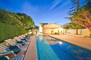 Une piscine avec des chaises longues, un espace de détente couvert et un bâtiment doté d’un toit en pente.