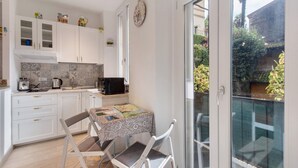 A compact kitchen with white cabinets, a dining table with a patterned tablecloth, and a view of a garden through a sliding glass door. - Studio Apartment 'Kella's Holiday Home' with Balcony, Wi-Fi and Air Conditioning (Bellagio)