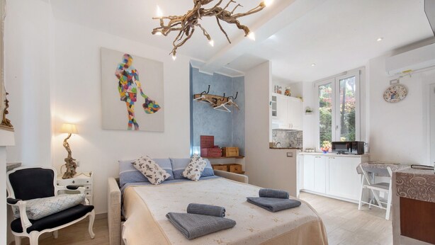 A bedroom with a bed, bedside tables, a chair, a desk, and a kitchen area. - Studio Apartment 'Kella's Holiday Home' with Balcony, Wi-Fi and Air Conditioning (Bellagio)
