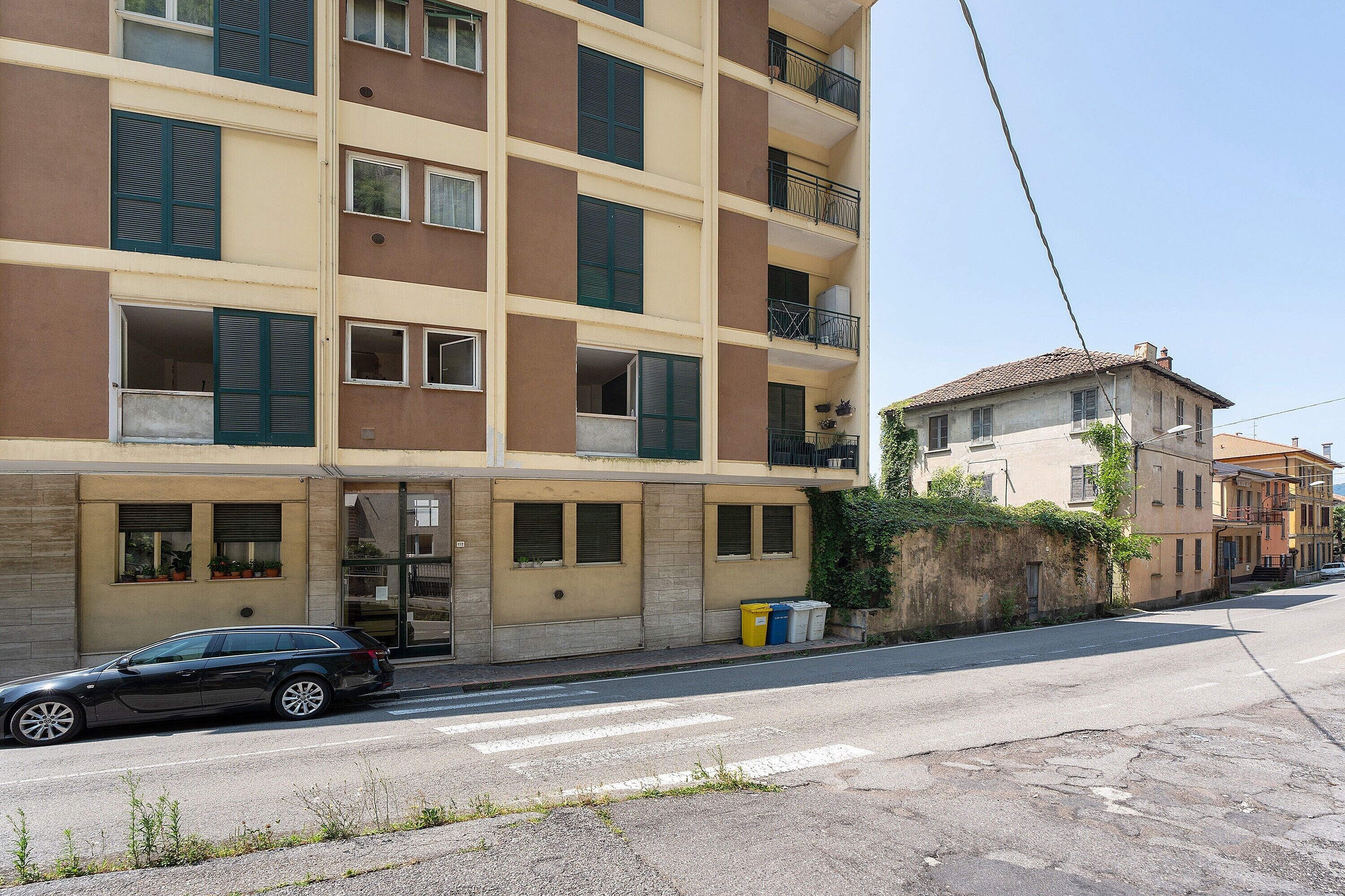 Un edificio residencial de varios pisos con balcones, un coche estacionado adelante y una calle con paso de peatones.