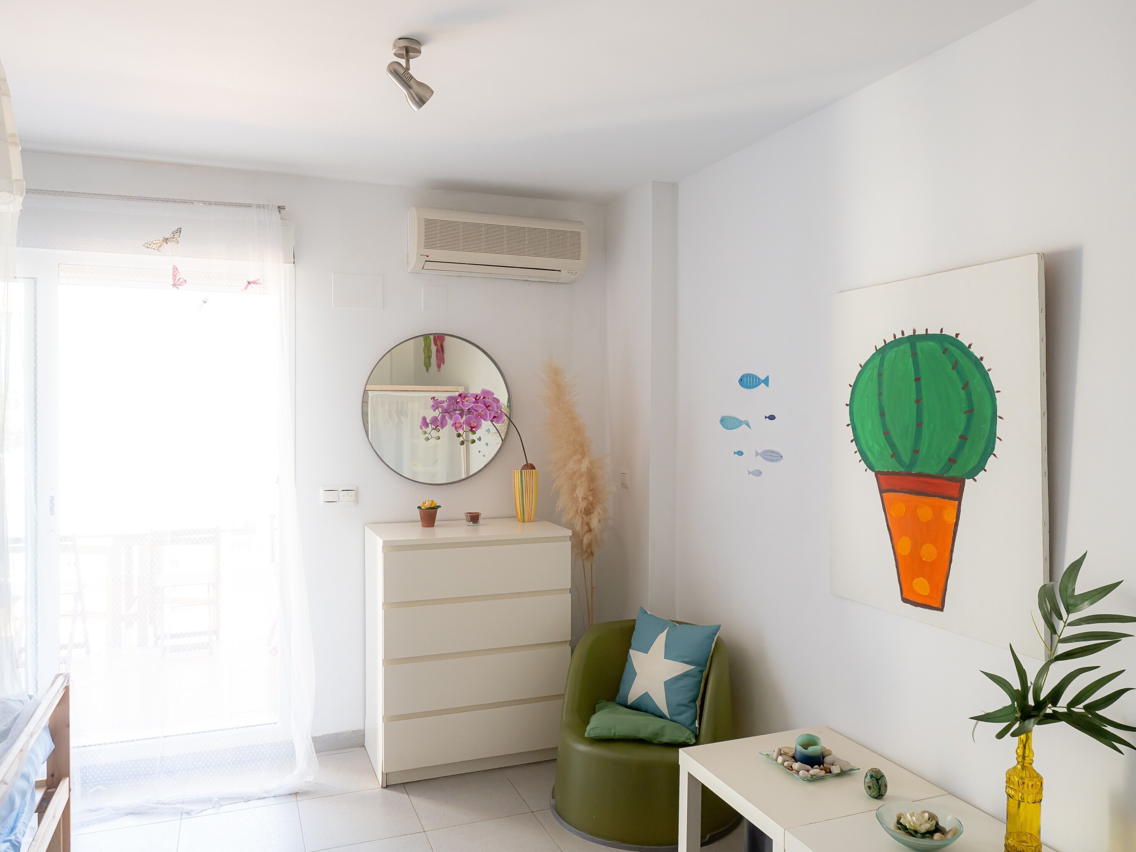 Une pièce avec une commode blanche, une chaise verte ornée d’un coussin en forme d’étoile, un miroir rond et un tableau représentant un cactus.