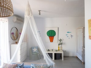 Une chambre d’enfant avec un lit, une table de chevet, un bureau, une chaise, un climatiseur fixé au mur, une fenêtre avec des rideaux et une décoration murale représentant un cactus.