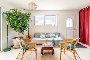 A living room with a sofa, two wicker chairs, a coffee table, a guitar, a large potted plant, and a window with a view.