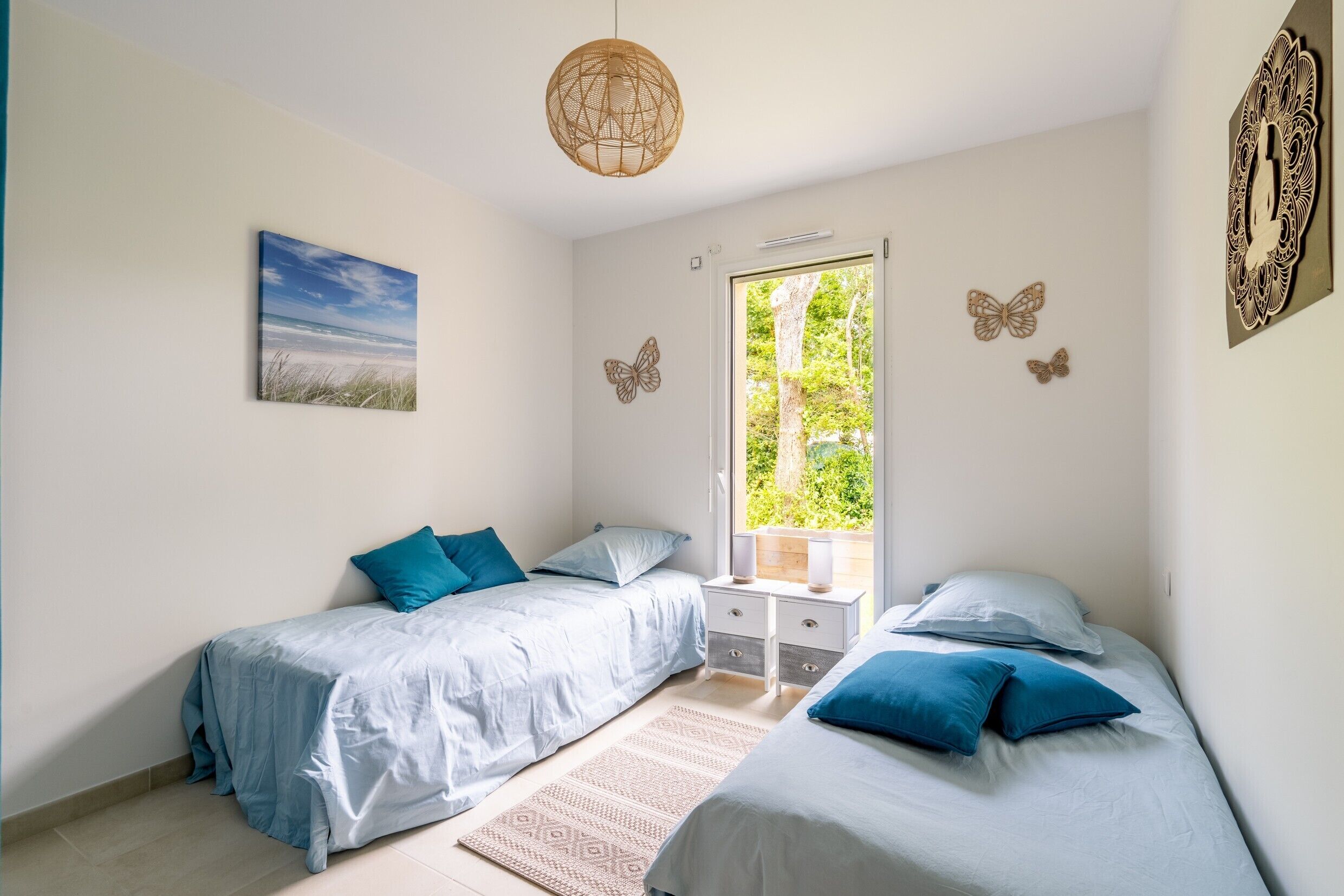 A bedroom with two beds, a window with a view of greenery, a decorative butterfly wall art, and a hanging light fixture.