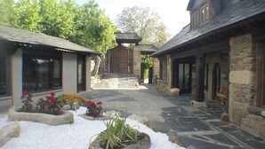 Un chemin de pierre mène à un bâtiment traditionnel doté d’un portail en bois, le tout étant entouré d’espaces aménagés avec des plantes et des rochers.