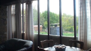 A room with a large window, a leather sofa, a dining table with chairs, and a basket of bread. - Country House 'Cuarto Del Ama' with Mountain Views, Private Garden and Wi-Fi (Rozas De Sanabria)