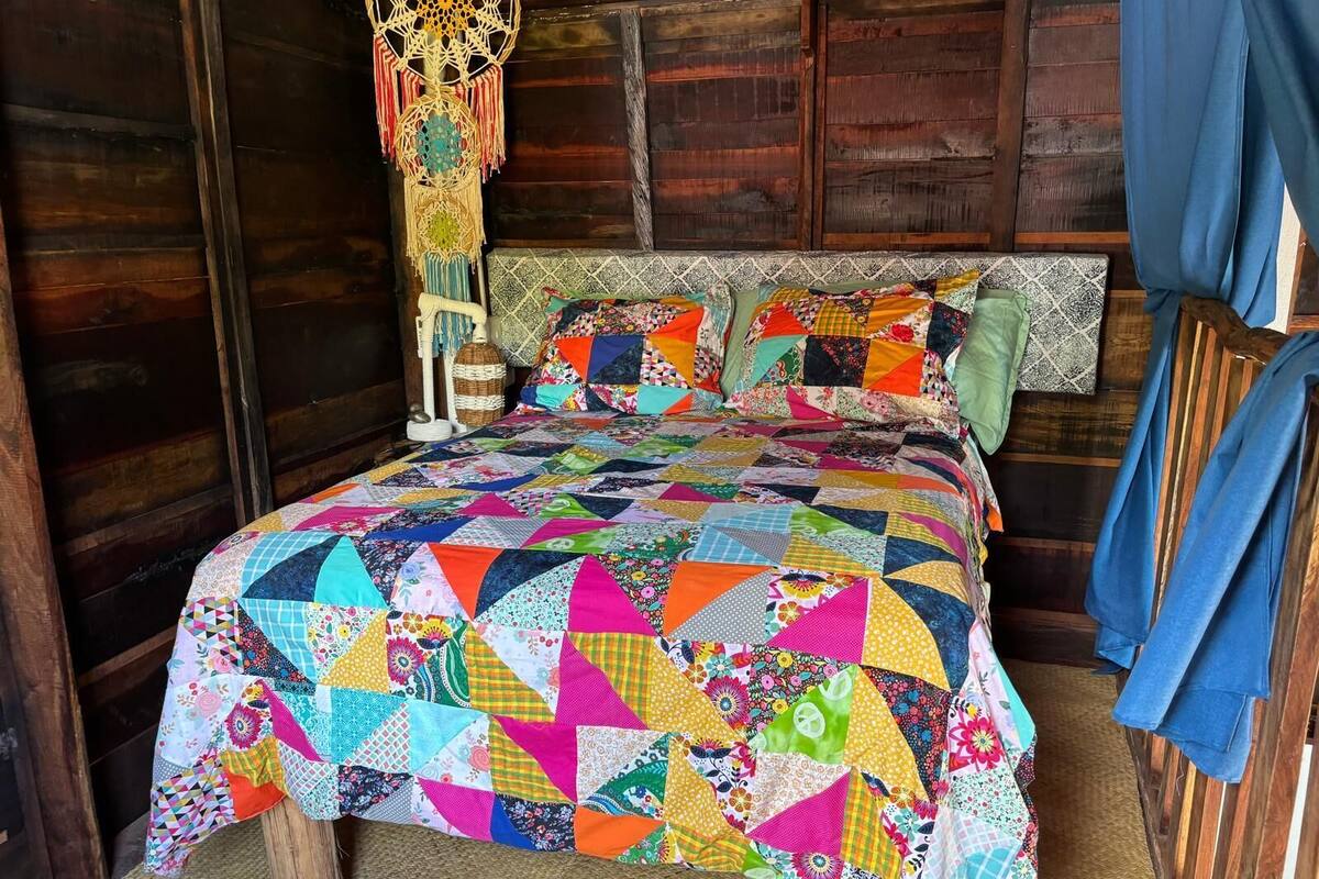 A cozy wooden room with a colorful patchwork bedspread, a woven hanging decoration, and a rustic headboard.