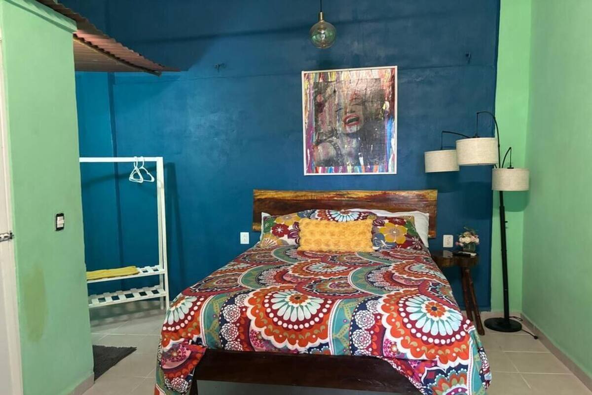 A bedroom with a colorful bedspread, a wooden headboard, a wall painting, and a bedside lamp.