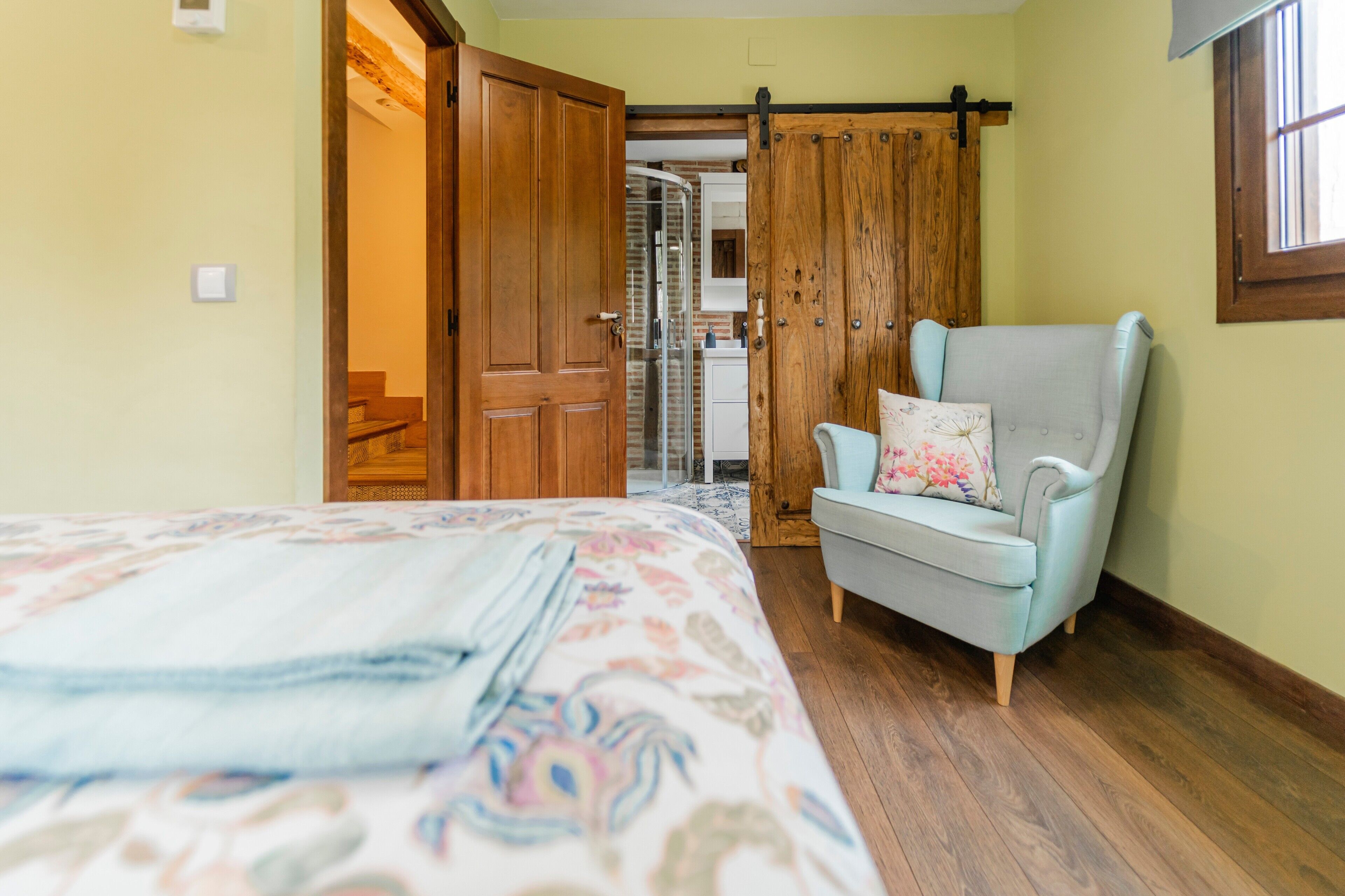 Una habitación con una cama, un sillón azul claro, una puerta de madera y una zona de ducha.
