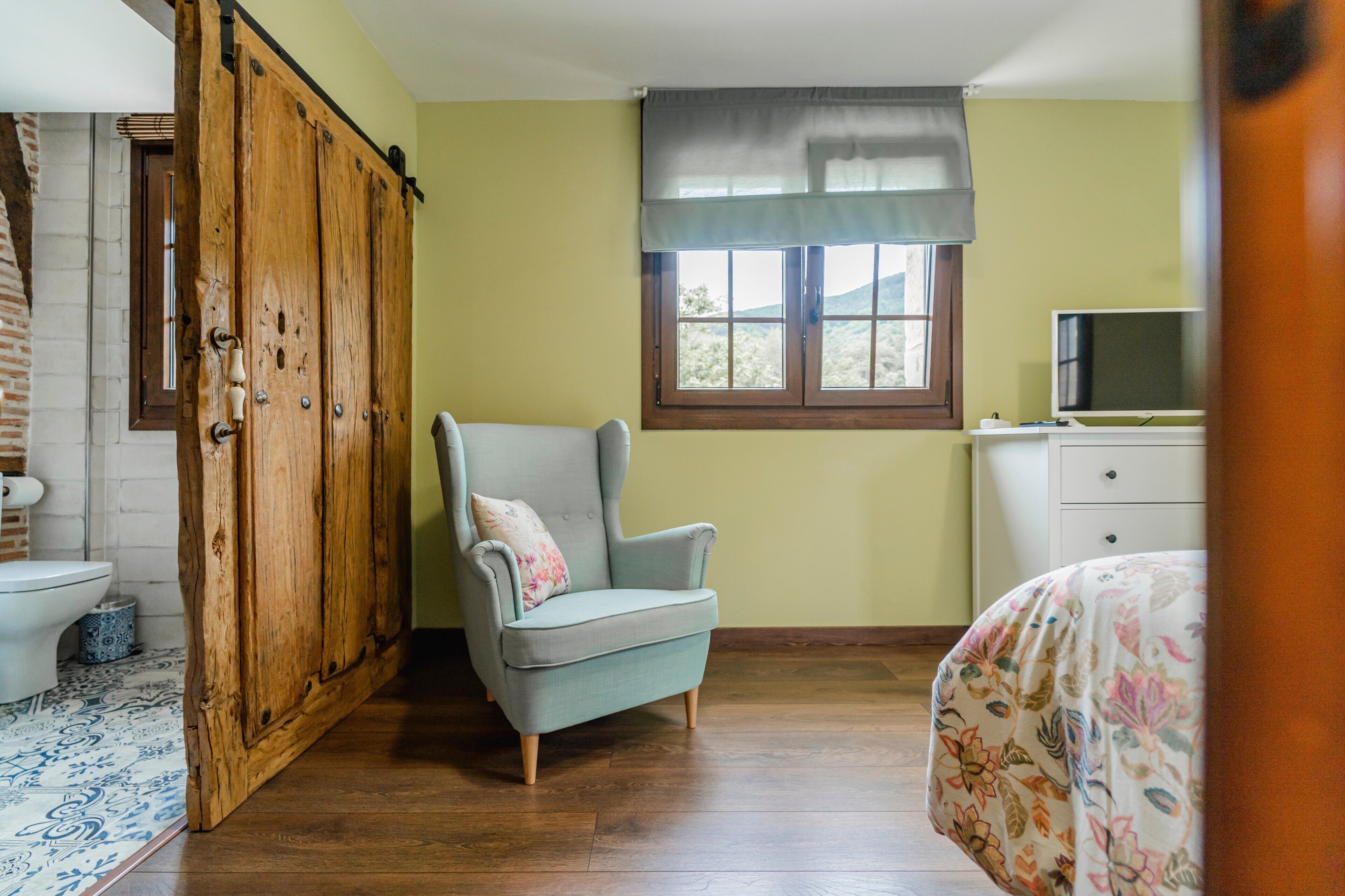 Une pièce comprenant une armoire en bois, un fauteuil gris, un lit recouvert d’une couvre-lit à motifs floraux, un téléviseur posé sur une commode et une fenêtre donnant sur un paysage verdoyant.