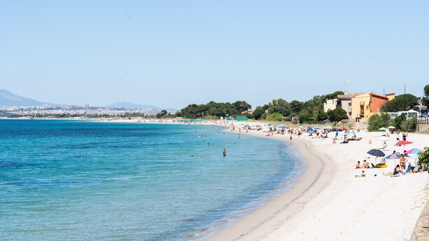 A beach with clear blue water, sandy shore, and buildings in the background.