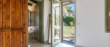 Puerta de madera abierta que da a un baño con lavamanos, inodoro y suelo de baldosas, con vistas a un jardín exterior.