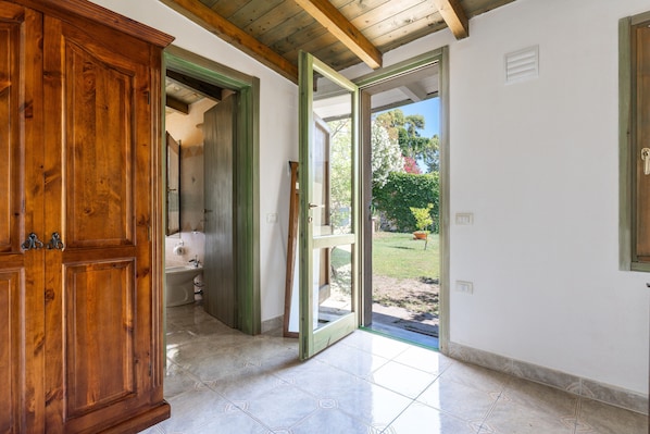 An open wooden door leading to a bathroom with a sink and toilet, a tiled floor, and a view of a garden outside.