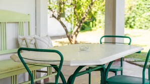 A green metal table and bench set with a white cushion, in a covered outdoor area with a view of greenery.