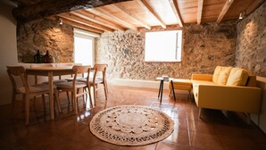 Una stanza accogliente con parete in pietra, travi in legno a soffitto, un divano giallo, un tappeto rotondo e di vimini e un tavolo da pranzo con sedie.
