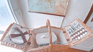 Un set da pranzo in vimini con piatti, posate e un piccolo contenitore, posizionato su un supporto bianco in una stanza con una finestra e una mappa appesa al muro.