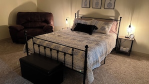 Un dormitorio con cama, una silla, una mesita de noche con lámpara y una pared decorada con obras enmarcadas.
