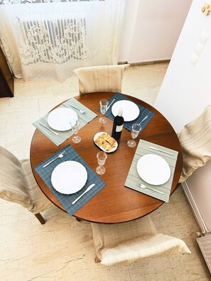 A round dining table with four plates, two wine glasses, a bottle of wine, and a small plate of food. - Sea Breeze Sanctuary-1bd 3pl 1ba - 90m To Sea (Chania)