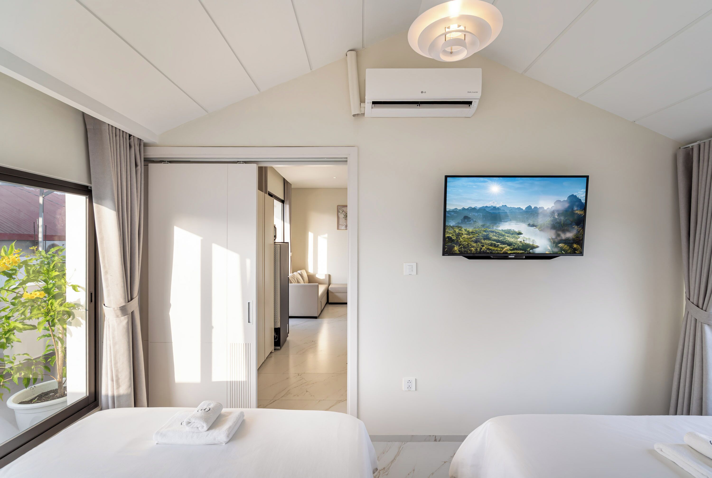 A hotel room with two beds, a TV mounted on the wall, and a potted plant by the window.