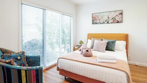 A bedroom with a large bed, a window with blinds, a chair with a colorful cushion, and a floral painting on the wall. - Modern Holiday House wth Bay View (Frankston South)