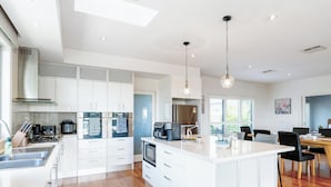 A modern kitchen with white cabinetry, a central island, and a dining area with a wooden table and chairs. - Modern Holiday House wth Bay View (Frankston South)