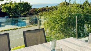 A dining area with a table, chairs, and a glass partition overlooking a swimming pool and greenery. - Modern Holiday House wth Bay View (Frankston South)