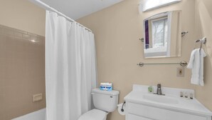 Un baño con inodoro, cortina de ducha, lavamanos con espejo y ventana con persianas.
