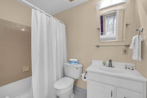 Une salle de bain comprenant des toilettes, un rideau de douche, un lavabo avec un miroir et une fenêtre avec des stores.