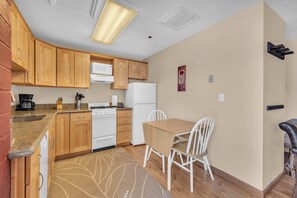 Cocina compacta con armarios de madera, una estufa blanca y un refrigerador. Hay una mesa de comedor con dos sillas y una alfombra estampada.