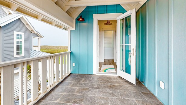 Ein überdachter Balkon mit Steinboden, Holzgeländer und einer Tür, die zu einem Flur mit blauer Wand und weißer Tür führt.