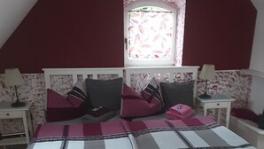 A bedroom with a sloped ceiling, a bed with a checkered bedspread, two nightstands with lamps, a window with floral curtains, and a wall sign.