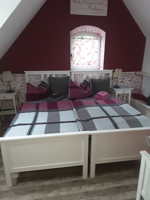 A bedroom with a sloped ceiling, a bed with a checkered bedspread, two nightstands with lamps, a window with floral curtains, and a wall sign.