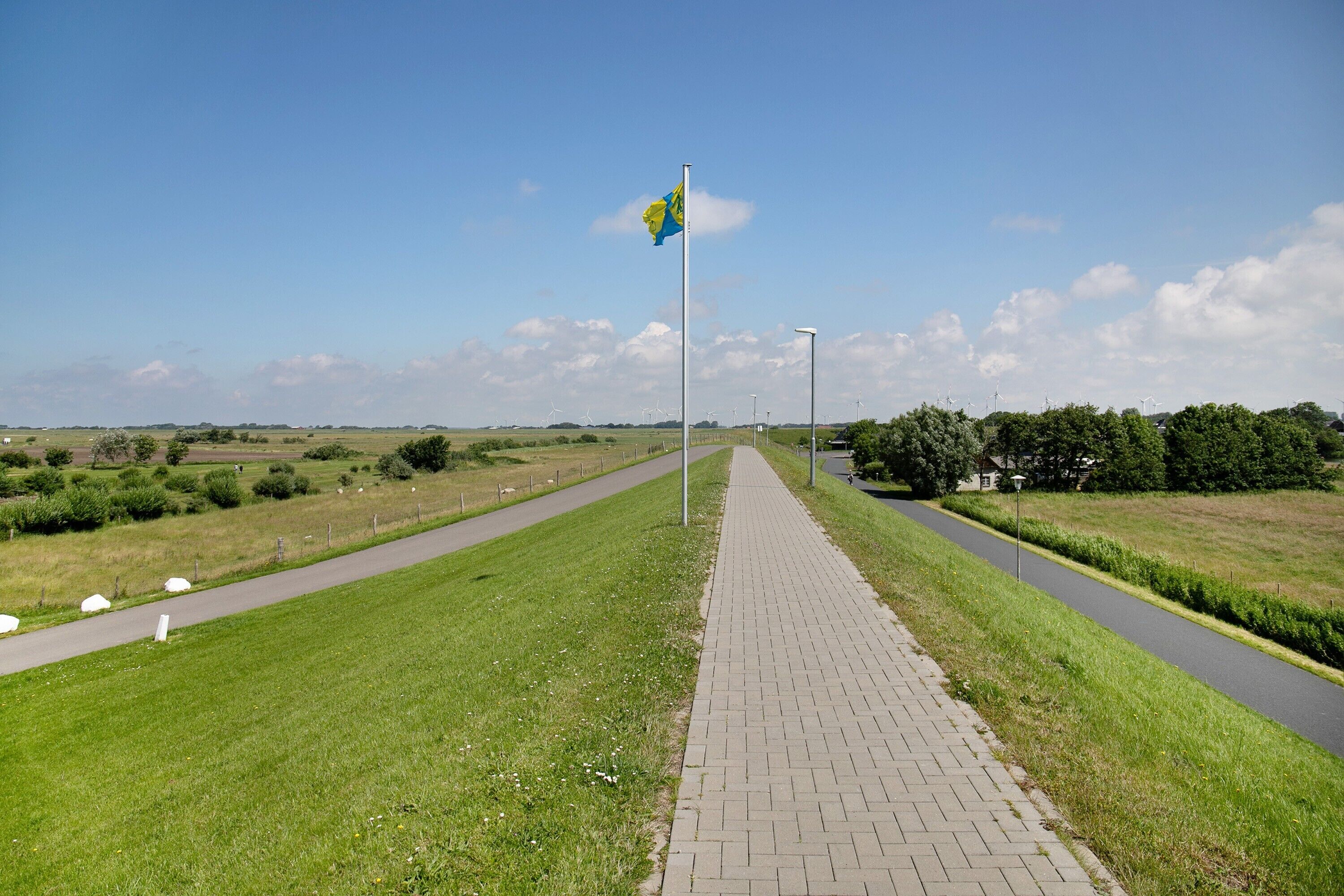 A paved pathway with a flagpole and flag, surrounded by grassy fields and wind turbines in the distance.