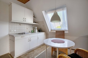 Une cuisine avec des meubles blancs, une table ronde surmontée d’un set de table violet, des chaises en osier et une fenêtre agrémentée d’un rideau blanc.