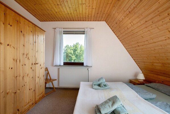 A room with a sloped ceiling, a bed with a pillow and a blanket, a radiator, a window with curtains, and a wooden wardrobe.