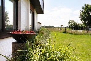 A white building with a window, a black planter with red flowers, and a grassy area with a fence in the background.