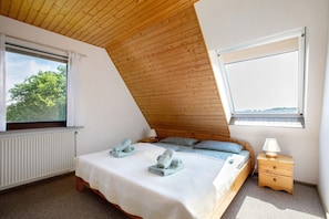 Une chambre avec un plafond en pente, un lit recouvert d’une courtepointe blanche, deux serviettes enroulées, une table de chevet avec une lampe et une fenêtre donnant sur les arbres.