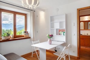 Une pièce avec une table blanche, deux chaises blanches, un vase avec des fleurs, une fenêtre donnant sur les montagnes et un meuble en bois.
