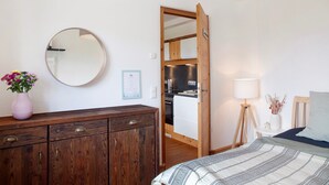 Un dormitorio con cómoda de madera, espejo redondo, cama con manta a rayas, mesita de noche con lámpara y vista a la cocina.