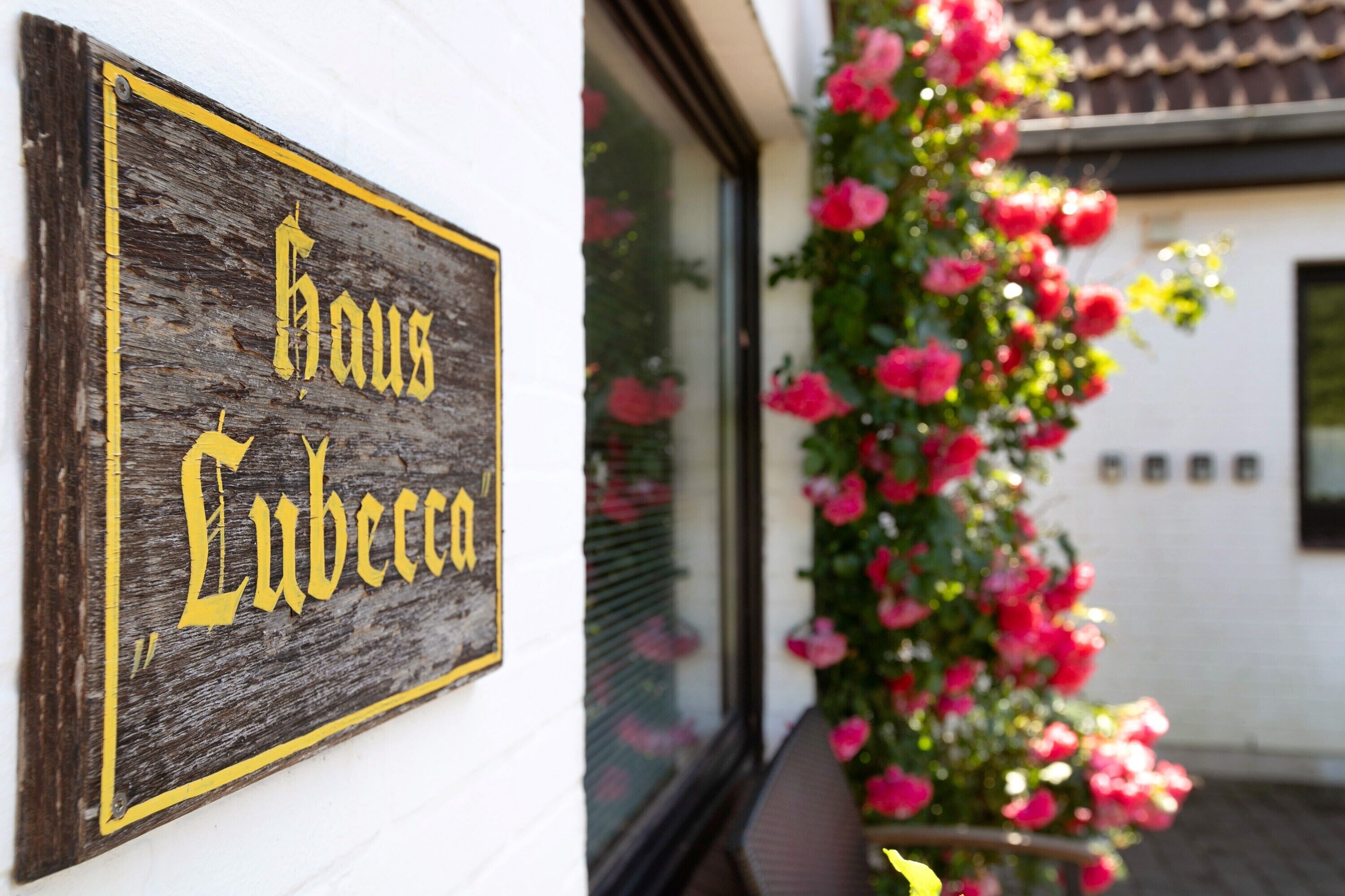 A wooden sign with 'Haus Lubeca' written on it, a white building with a tiled roof, and a potted plant with pink flowers.