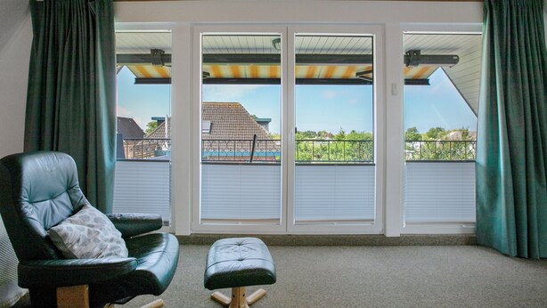 A room with a black leather chair, a wooden ottoman, and a view through large sliding glass doors to a balcony with a metal railing. - Apartment 'Balkonwohnung' with Shared Garden, Balcony and Wi-Fi (Westerdeichstrich)