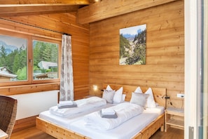 A wooden cabin bedroom with two beds, a window with curtains, a wall-mounted picture, and a nightstand.