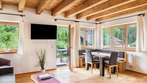 A spacious room with wooden beams, a dining area with a table and chairs, a flat-screen TV mounted on the wall, and large windows offering a view of greenery outside.