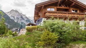 A wooden chalet with a balcony, surrounded by lush greenery and mountains in the background.
