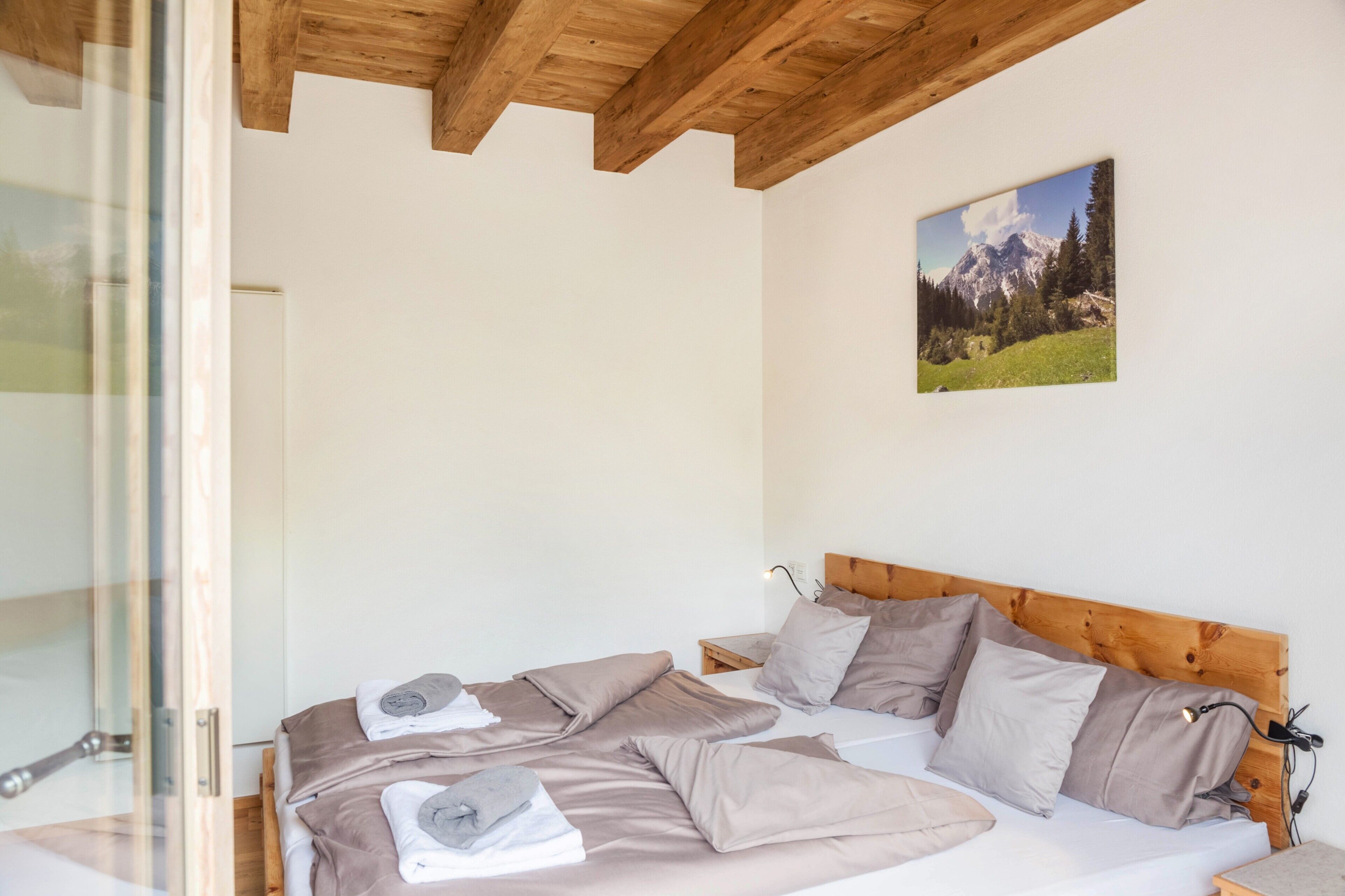 A bedroom with a wooden ceiling, a bed with multiple pillows, a nightstand, and a painting of a mountain landscape.